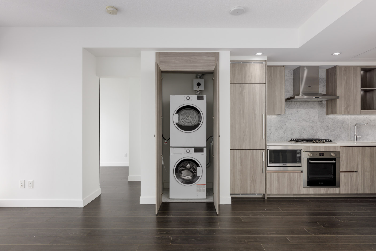 A modern kitchen with built-in light wood cabinets, stainless steel appliances, and a stacked washer and dryer in a closet with open doors.