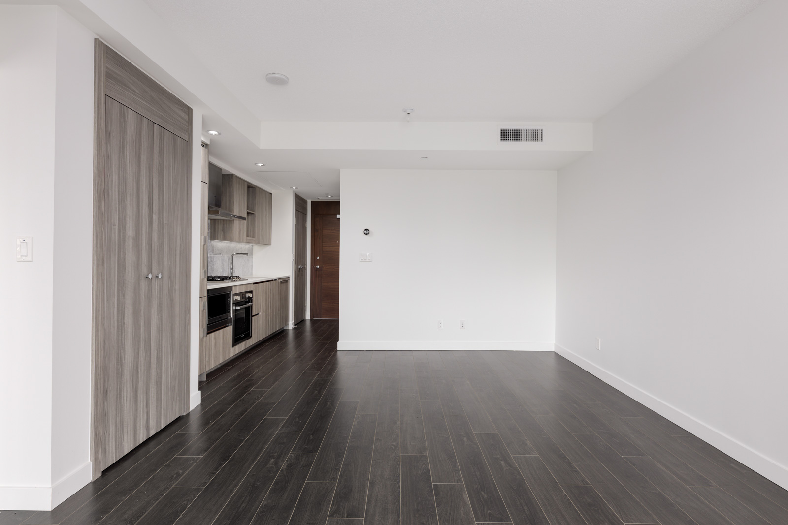 Unfurnished modern apartment interior with dark wood flooring, white walls, built-in kitchen along the left, and a closed closet door near the entrance.