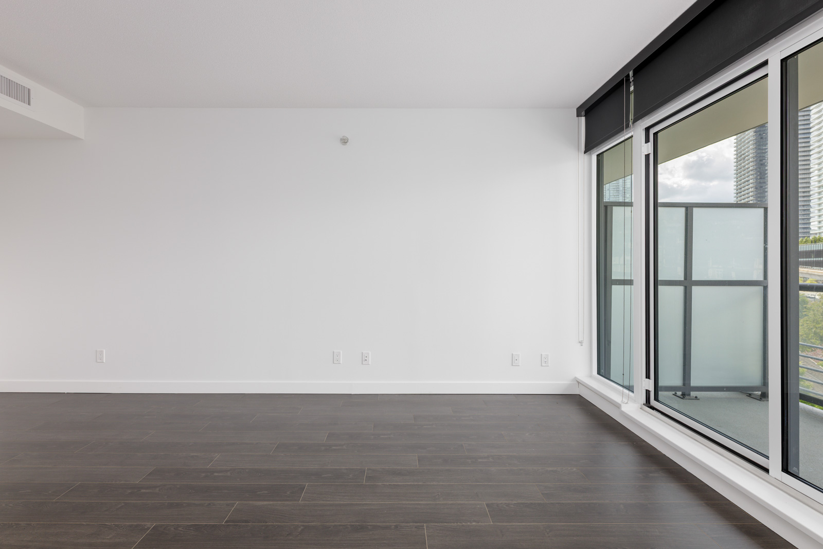 Empty room with white walls, dark wood flooring, large windows, and a glass balcony door showing an outdoor view.