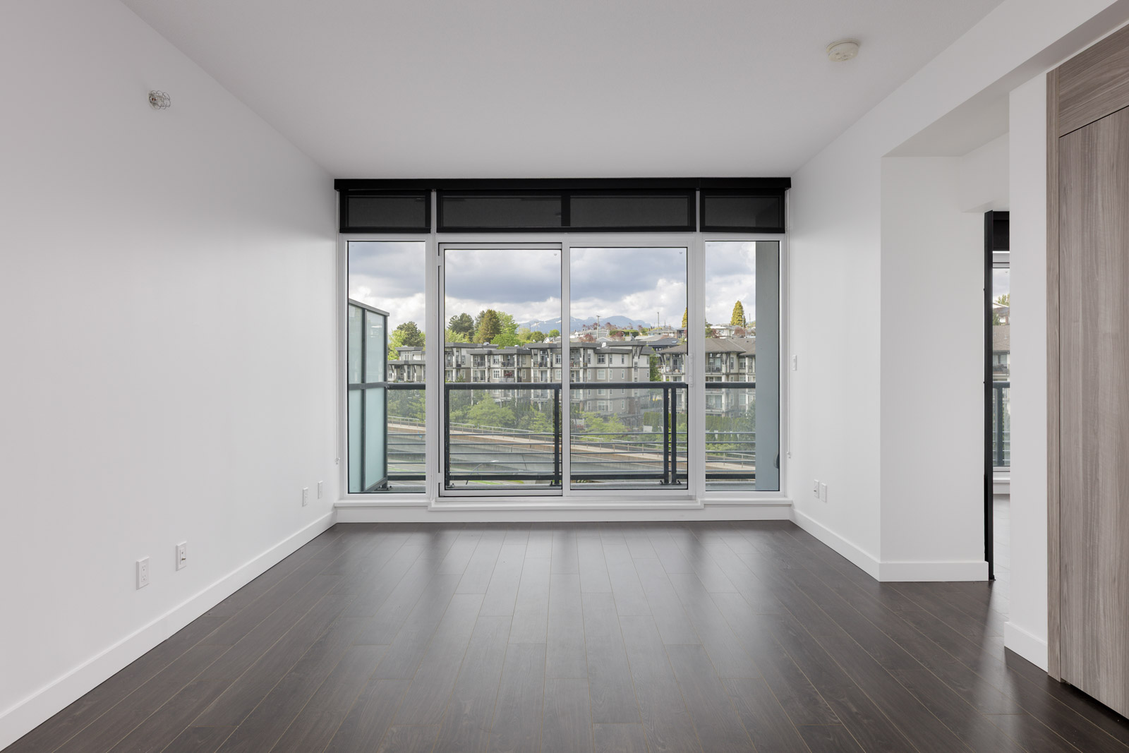 Unfurnished apartment room with dark wood flooring, white walls, and large floor-to-ceiling windows leading to a balcony with a view of nearby buildings and greenery.