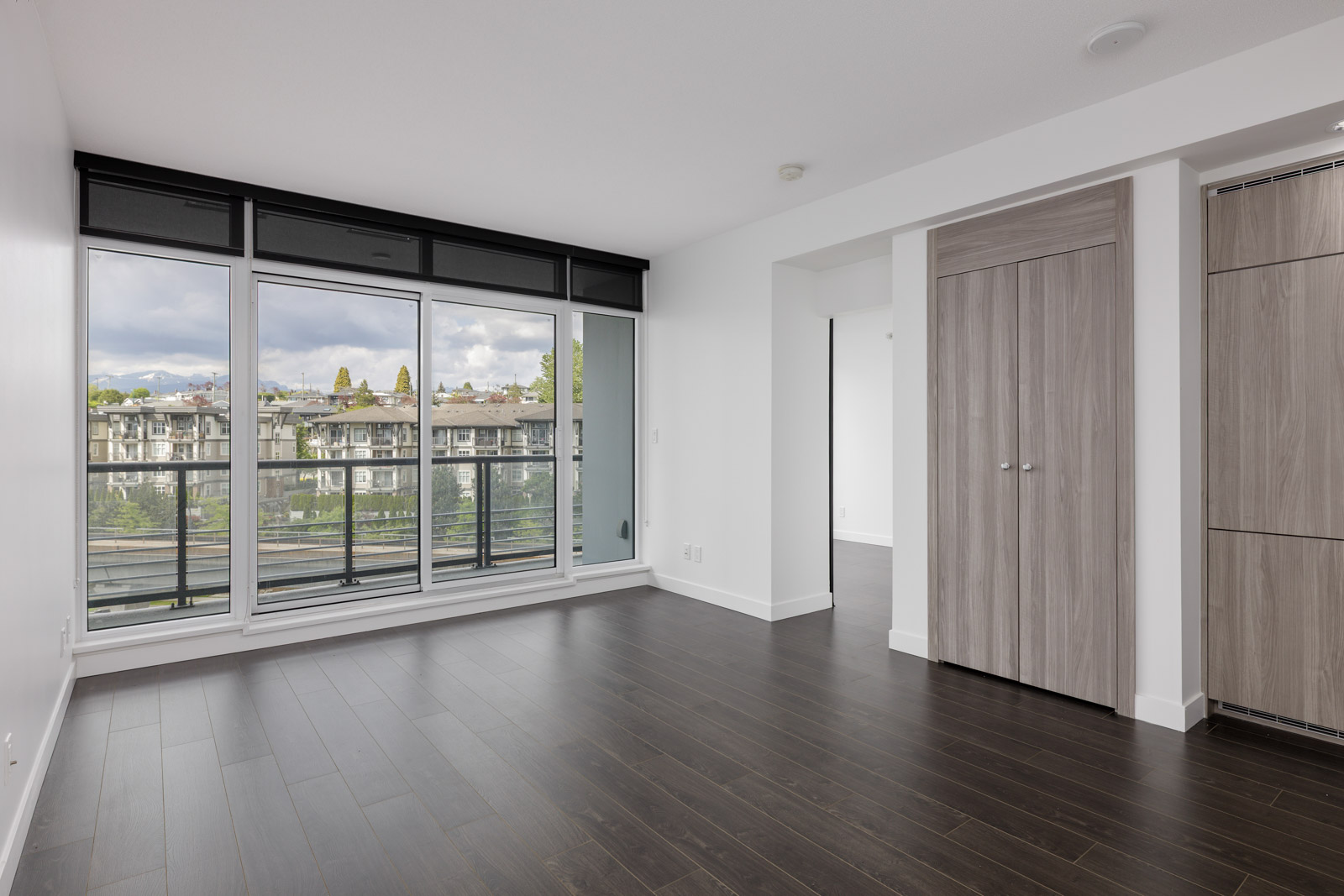 Unfurnished modern living room with dark wood floors, large windows leading to a balcony, and a closed light wood closet.