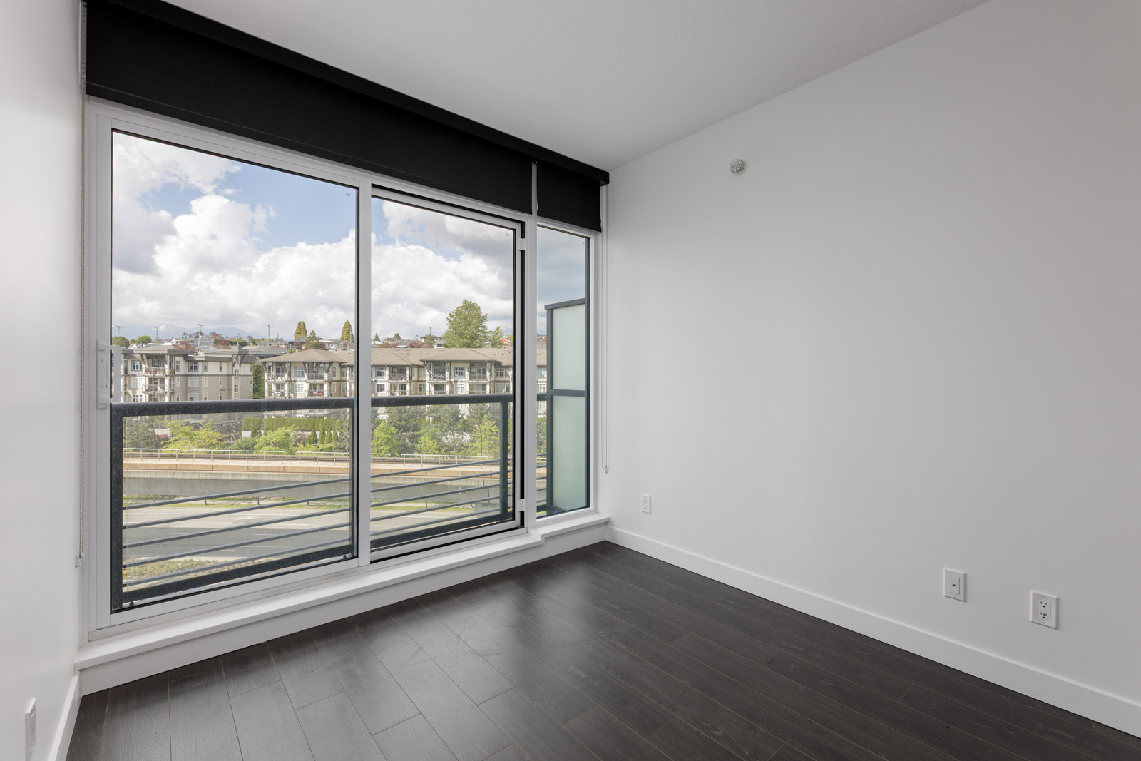 Empty room with dark wood flooring, white walls, and large floor-to-ceiling windows overlooking a balcony and neighboring buildings under a partly cloudy sky.