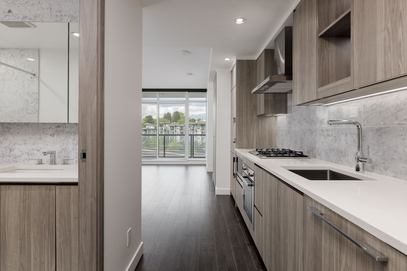 Modern kitchen with light wood cabinets, white countertops, and built-in appliances adjacent to a bathroom; large windows provide natural light to the open living area.