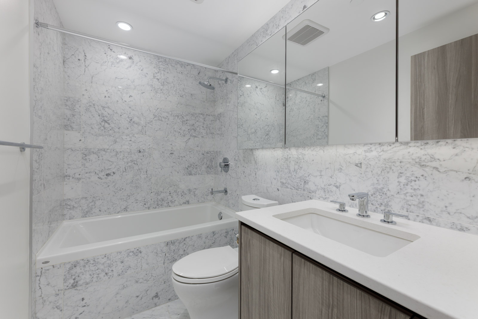 Modern bathroom with marble walls, a bathtub-shower combo, toilet, large mirror, and a sink with a white countertop and wood cabinetry.