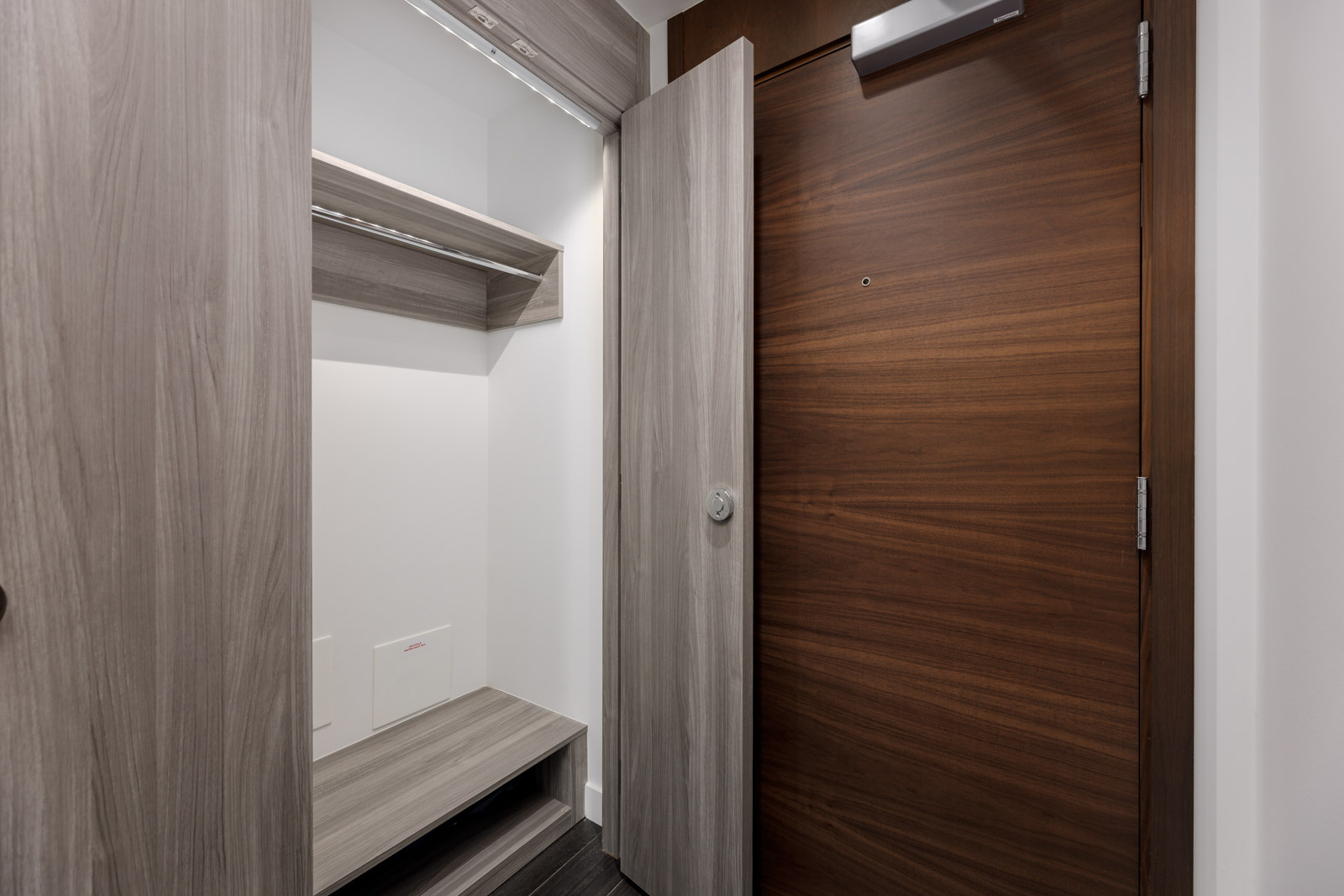 A modern entryway with a wooden door, a built-in closet featuring shelves and a bench, and gray wood paneling.