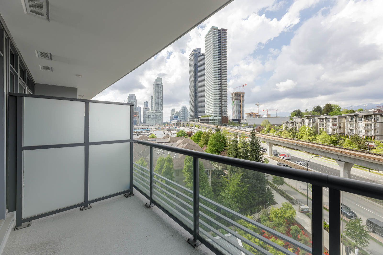 A modern balcony with glass railing overlooks a cityscape with tall buildings, a highway, and trees under a partly cloudy sky.
