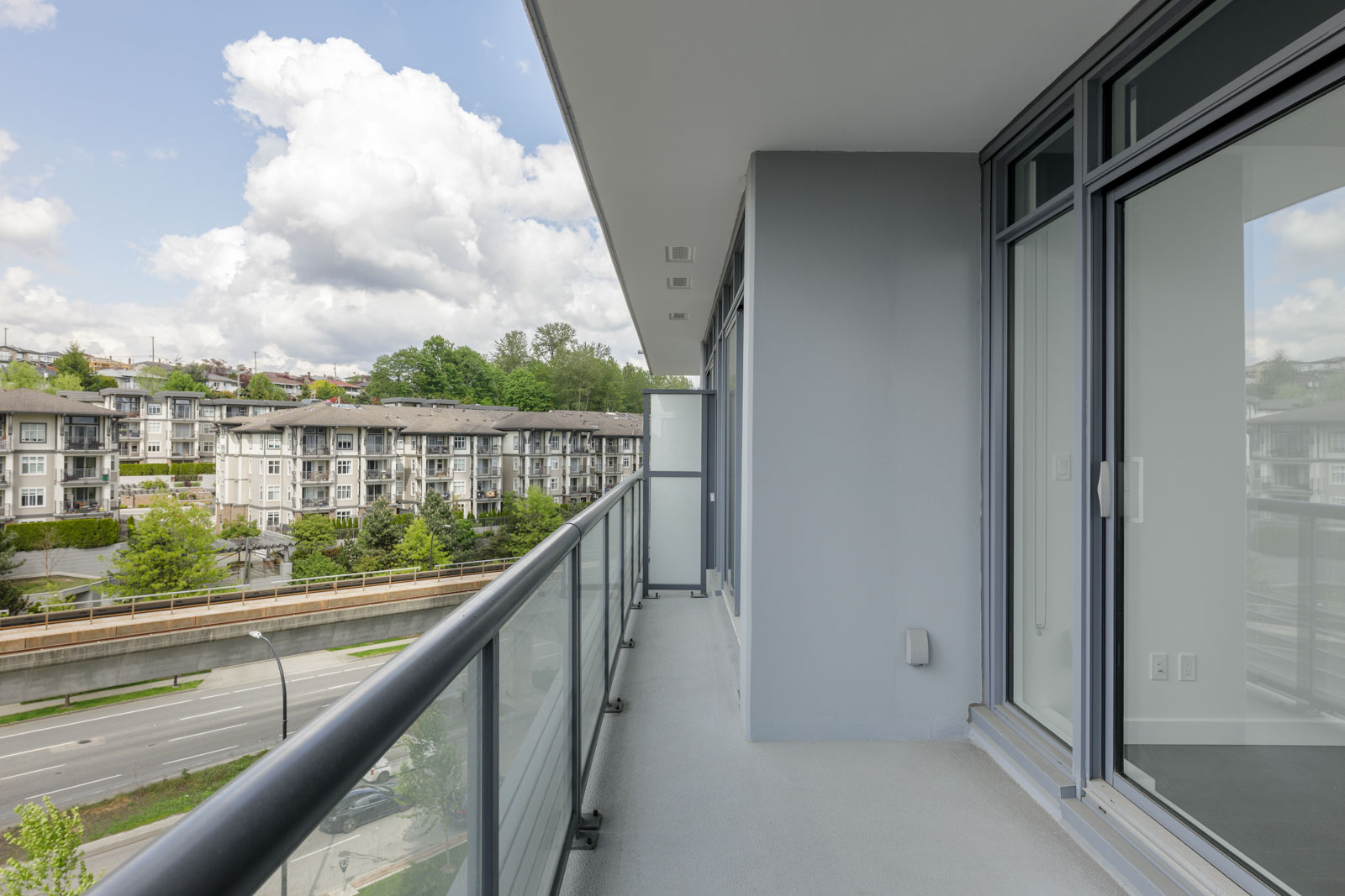 A modern apartment balcony with glass railings overlooks a street and residential buildings, with trees and a cloudy sky in the background.