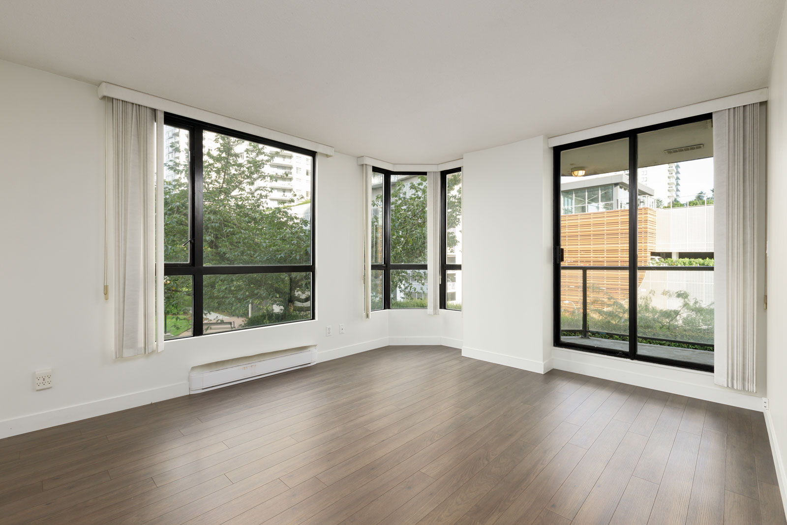 Empty corner room with large floor-to-ceiling windows, white walls, and dark wood flooring, overlooking trees and modern buildings outside.