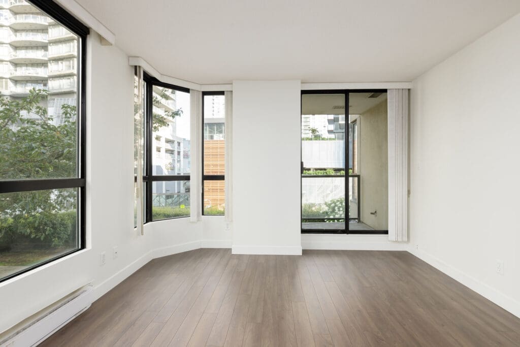 Empty modern room with large windows, white walls, and wood flooring, overlooking greenery and nearby buildings.