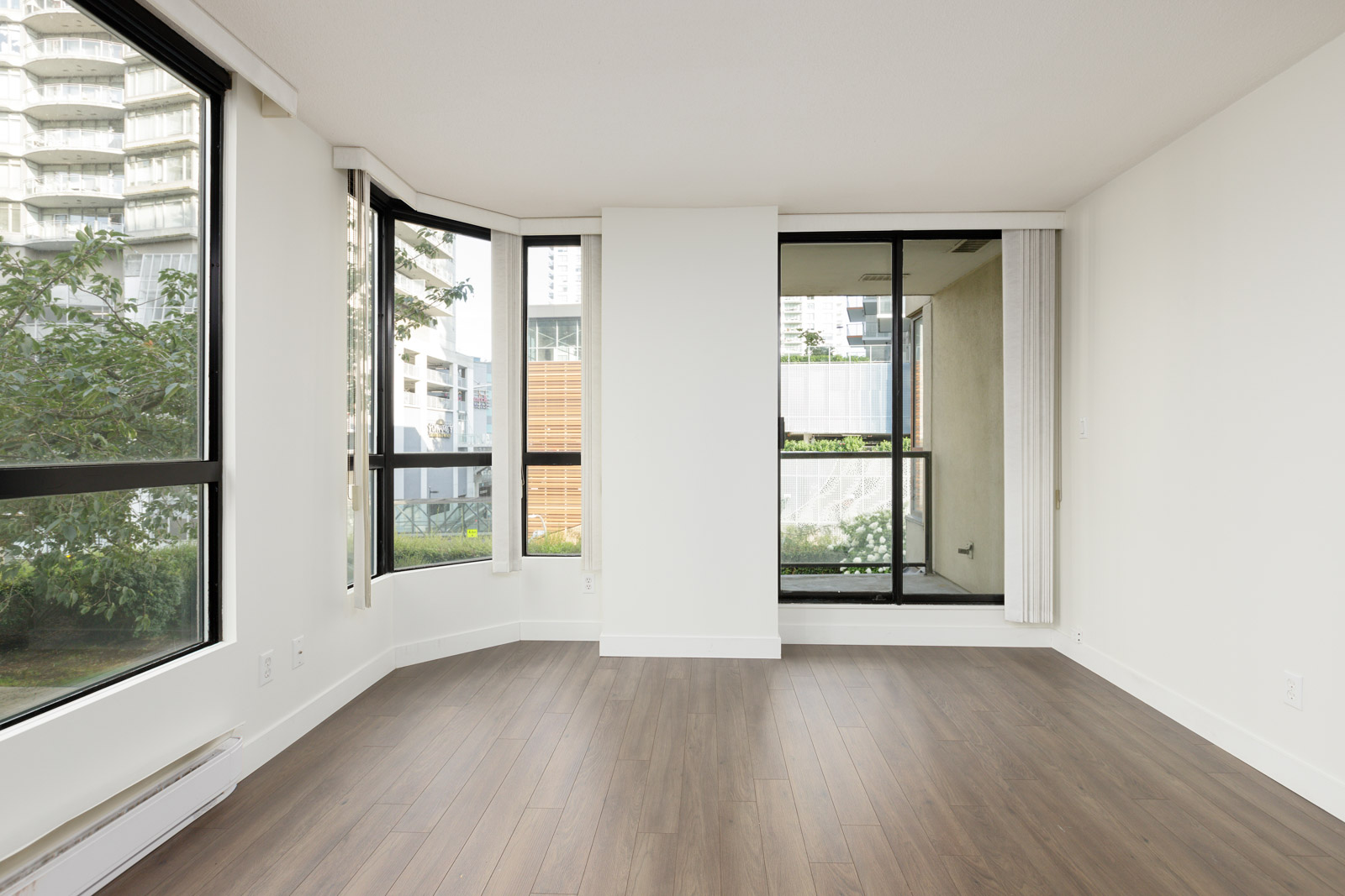 Empty modern room with large windows, white walls, and wood flooring, overlooking greenery and nearby buildings.