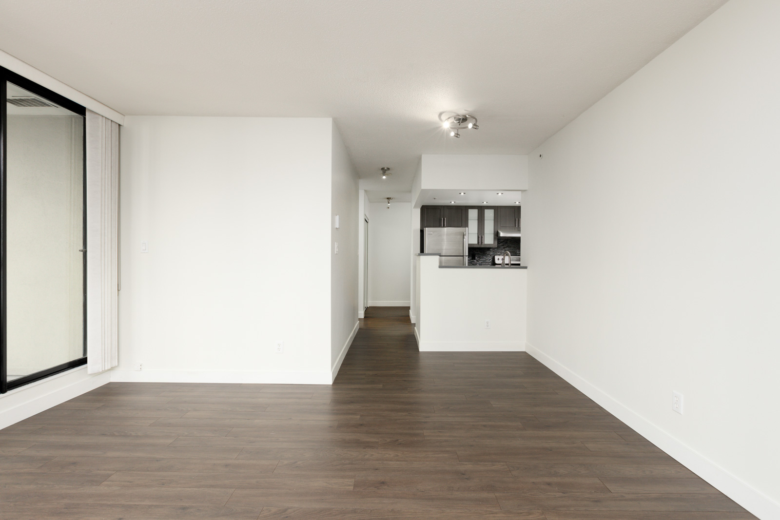 Unfurnished apartment with white walls, large window, dark wood floors, and an open view into a modern kitchen with stainless steel appliances.