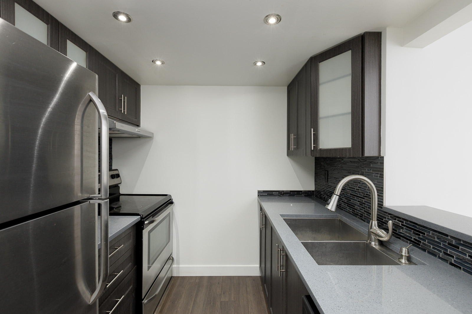 Modern kitchen with stainless steel appliances, dark cabinets, quartz countertops, double sink, and tile backsplash under bright recessed lighting.