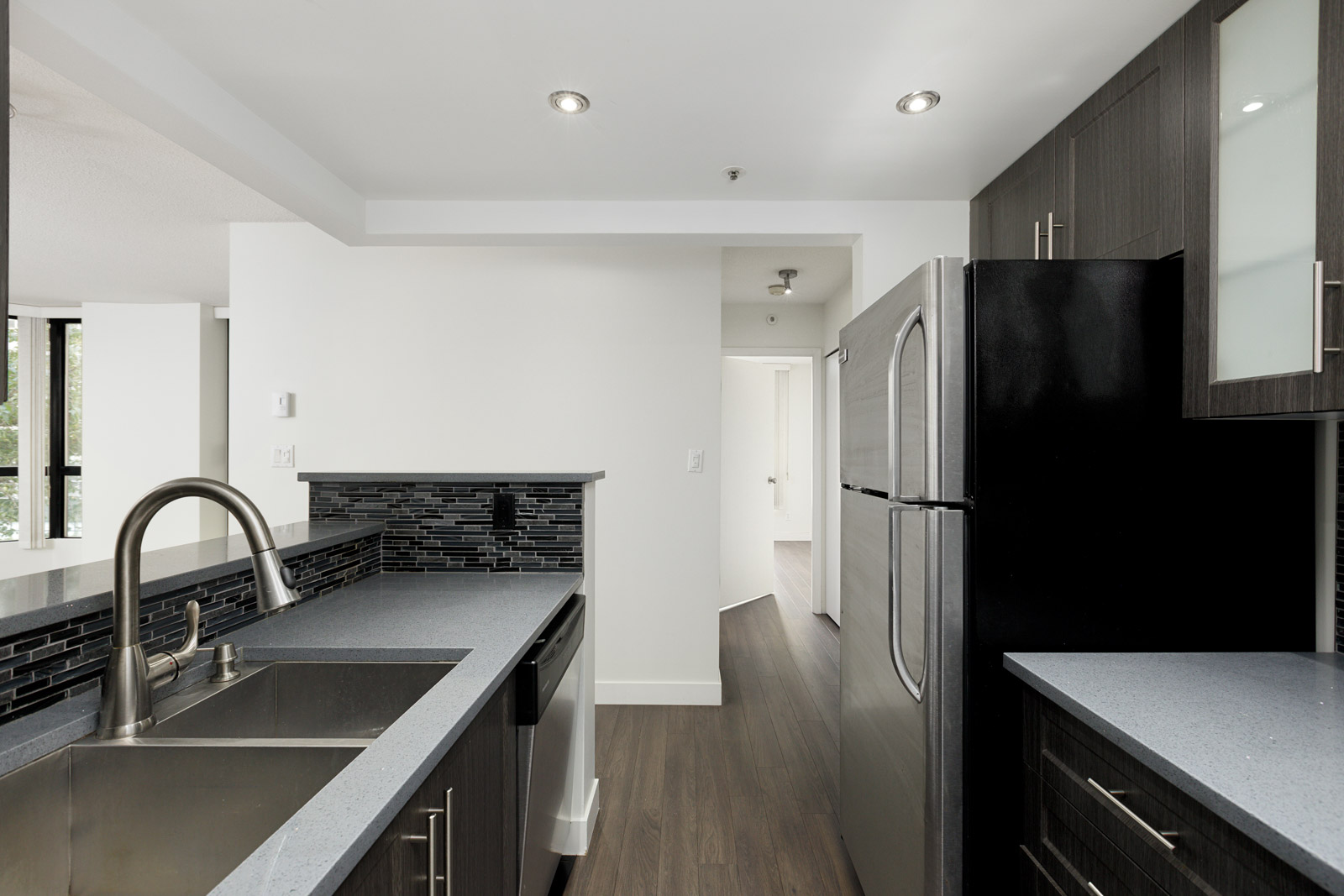 Modern kitchen with dark cabinets, stainless steel appliances, double sink, and light gray countertops, featuring a tile backsplash and wood flooring.