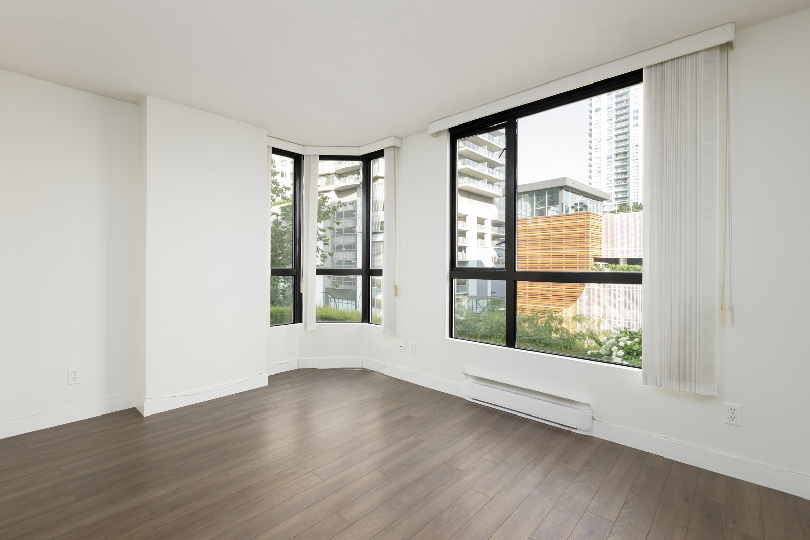 Empty room with large corner windows, white walls, and wood flooring, overlooking modern buildings and greenery outside.