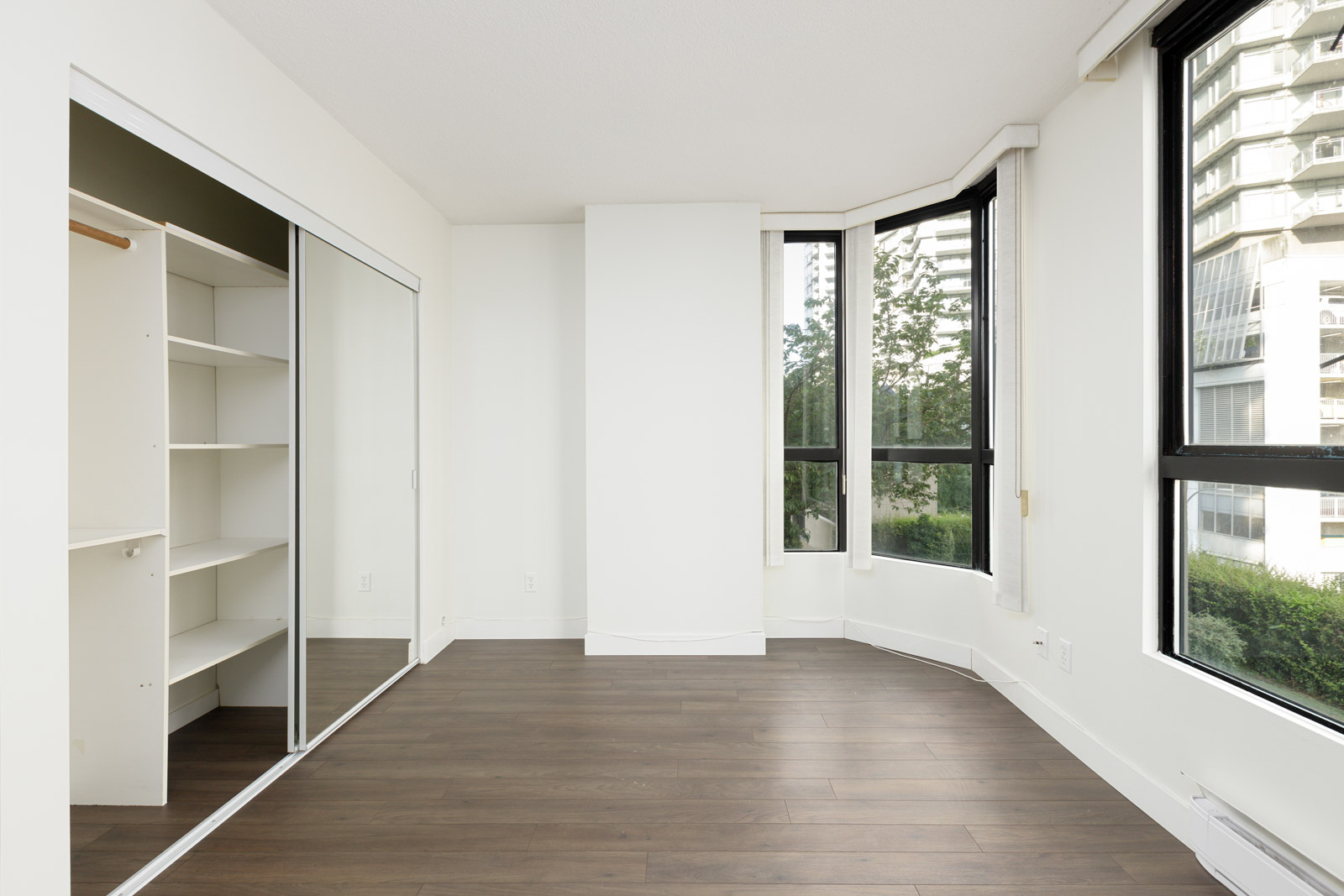 Empty room with large windows, white walls, dark wood flooring, and an open closet with shelves and a mirrored sliding door.