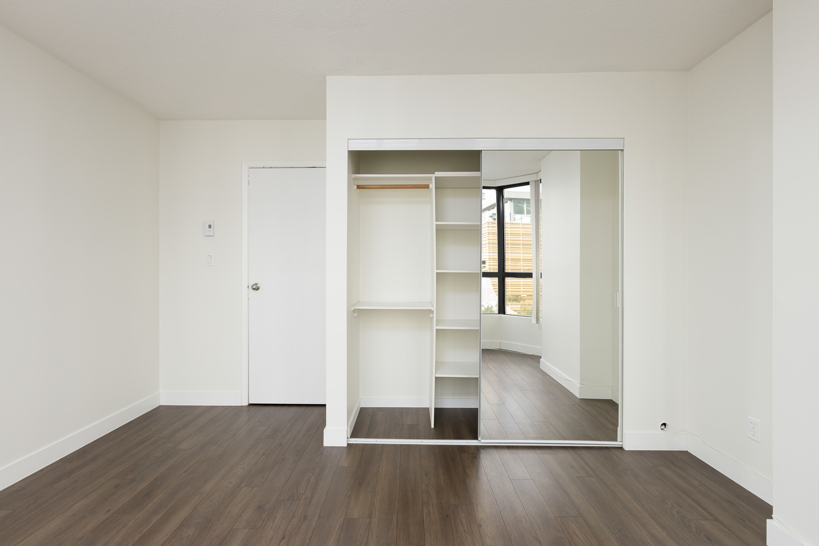 Empty room with white walls, wood flooring, a closet with sliding mirrored doors, and a window in the background letting in natural light.