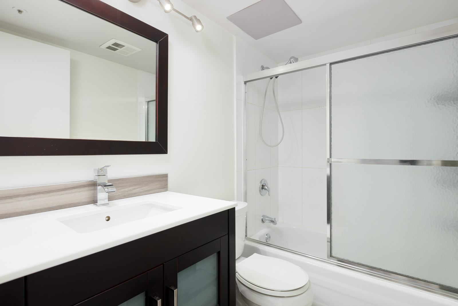 Modern bathroom with a vanity sink, large mirror, toilet, and a bathtub with sliding glass doors and a handheld showerhead.