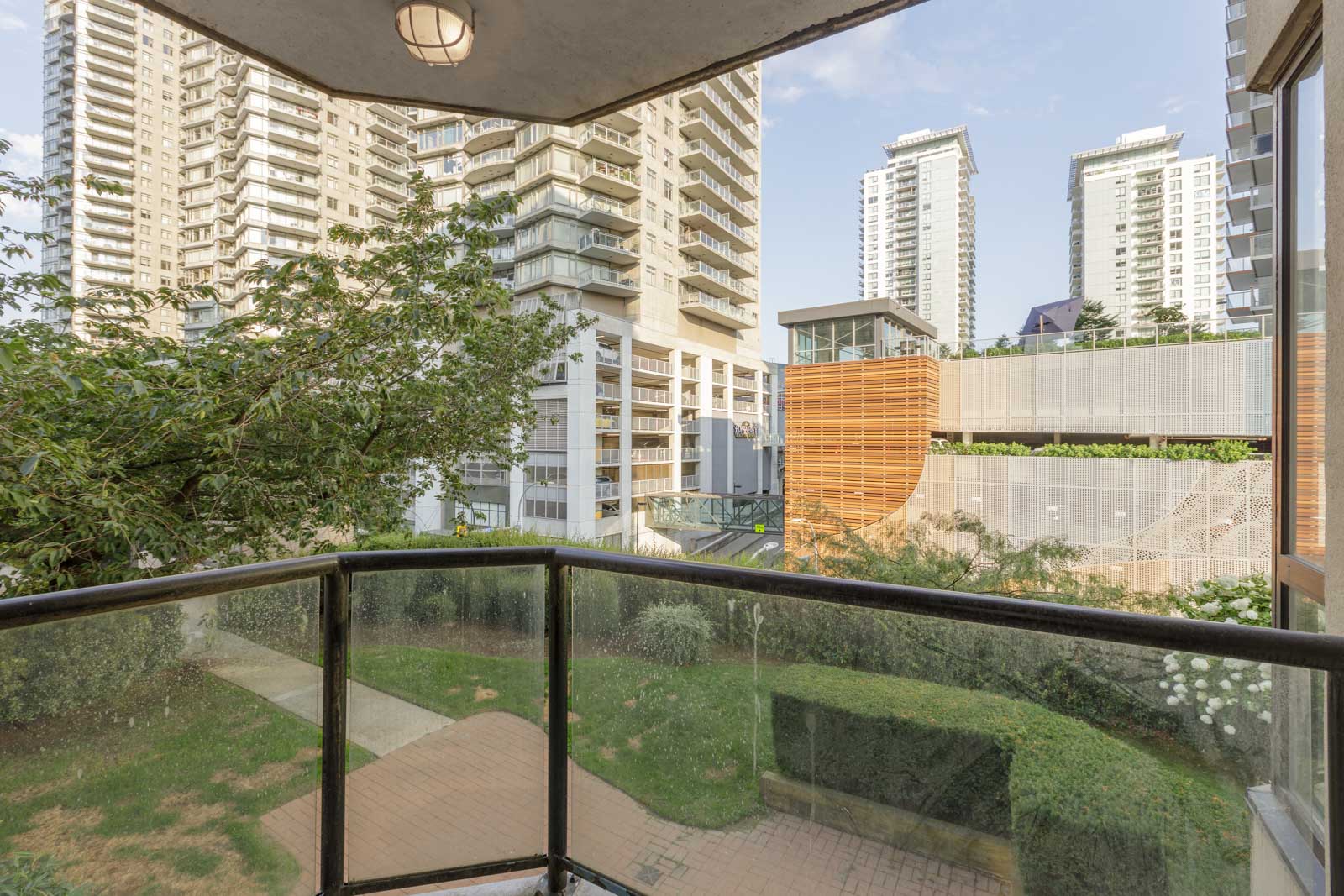 View from a balcony with glass railing overlooking a courtyard, landscaped greenery, and several modern high-rise buildings.
