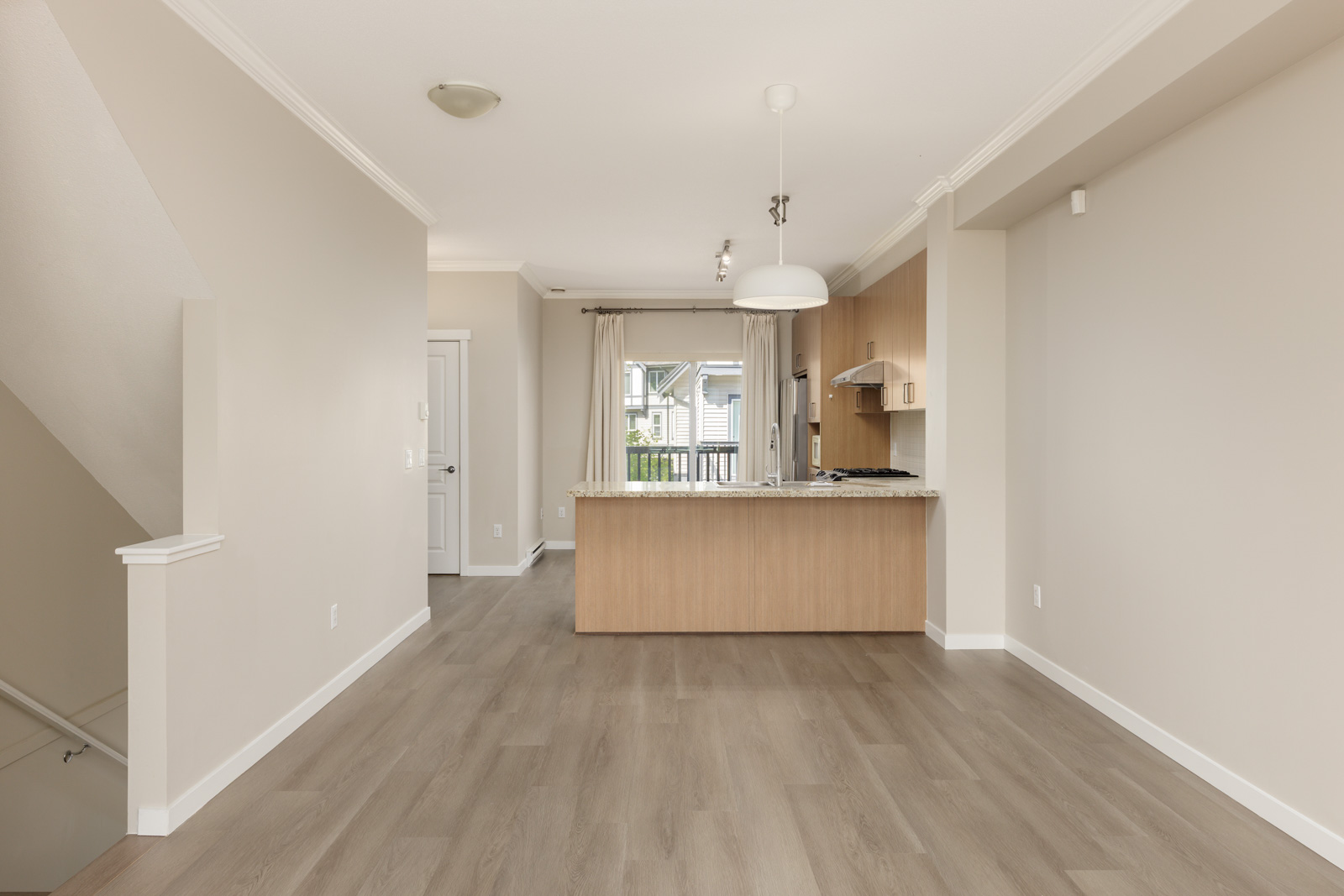 Unfurnished modern kitchen and dining area with light wood floors, beige walls, and a staircase on the left. Natural light enters through a window behind the kitchen.