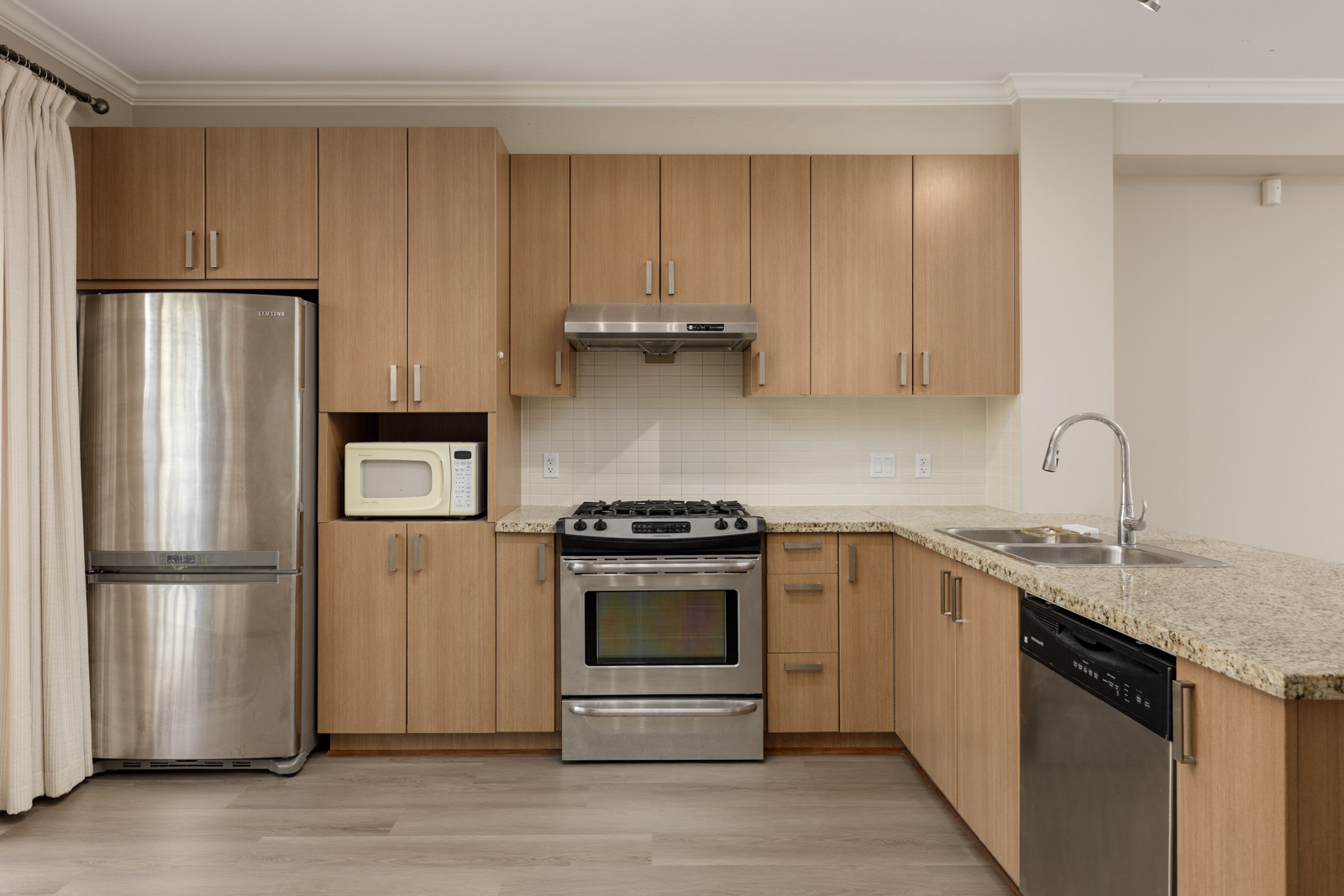 Modern kitchen with wooden cabinets, stainless steel appliances including a fridge, gas stove, oven, dishwasher, microwave, and granite countertops with a double sink.