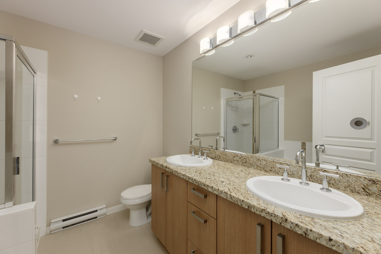 Modern bathroom with double sinks, granite countertop, large mirror, toilet, towel bar, and glass shower enclosure. Walls are beige and lighting is above the mirror.