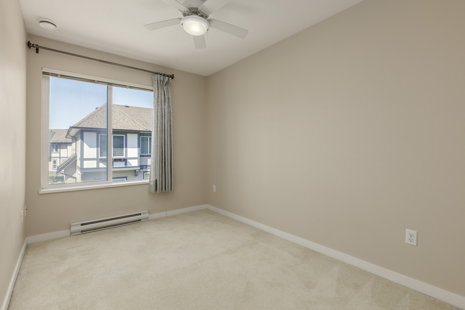 Empty beige-walled room with light carpet, ceiling fan, single window with curtain, and view of neighboring building outside.
