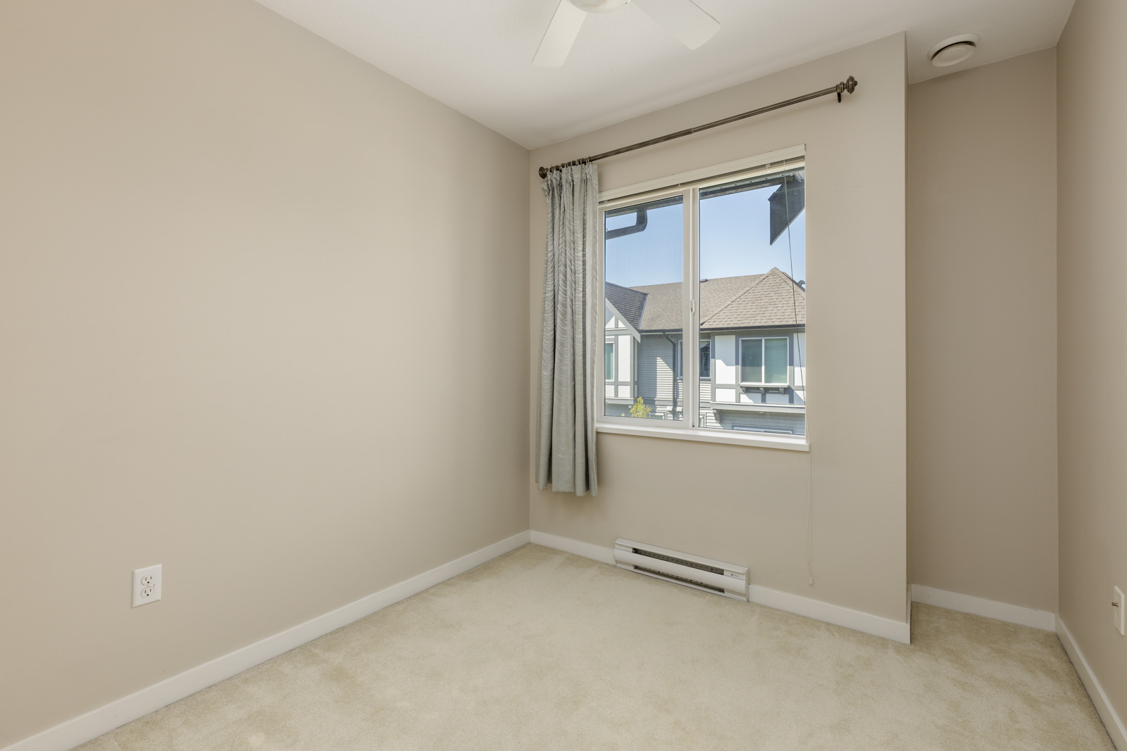 Empty beige room with cream carpet, a window with gray curtains, ceiling fan, and view of neighboring houses outside the window.