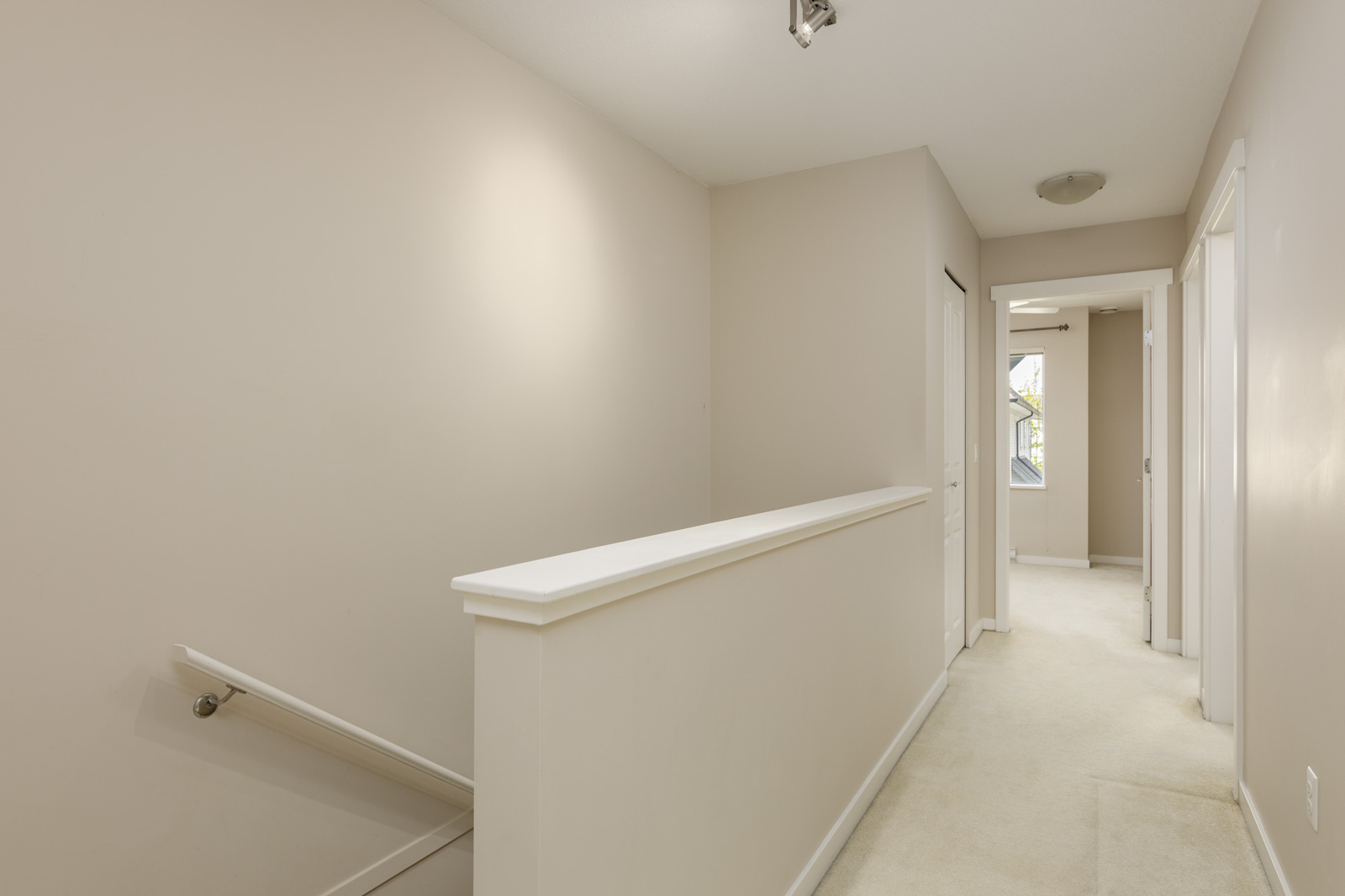 A hallway with beige walls and carpet, featuring a stairway on the left and doors leading to separate rooms on the right and at the end.