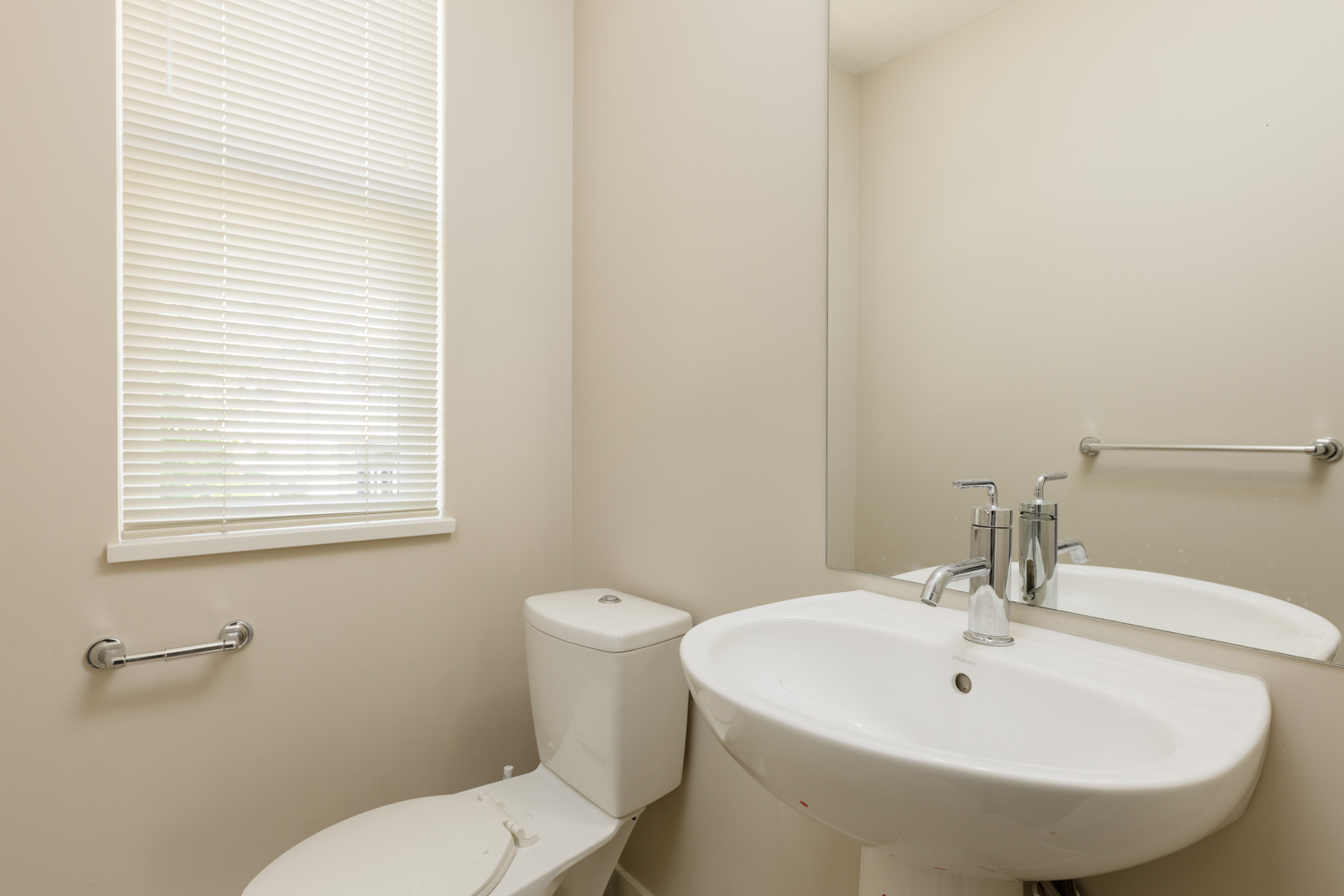 A small bathroom with a toilet, a white sink, a large wall mirror, and a window with closed blinds.