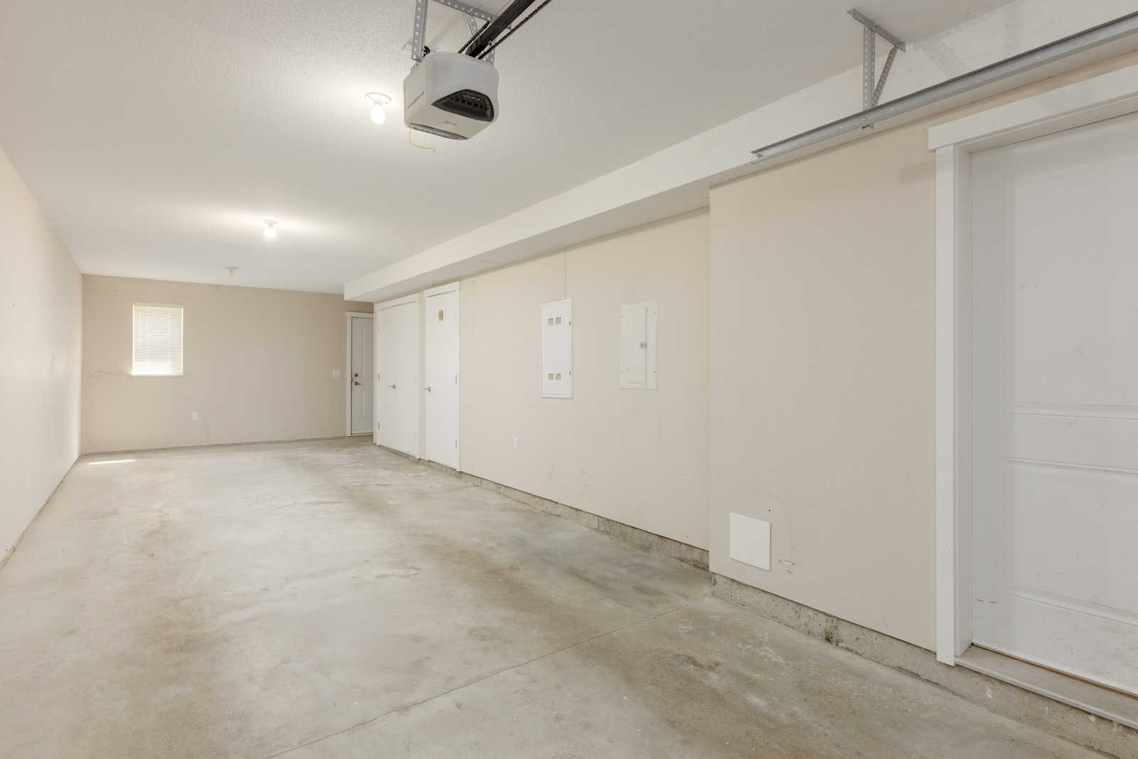 Empty two-car garage with beige walls, exposed concrete floor, ceiling lights, electrical panels, and multiple doors.