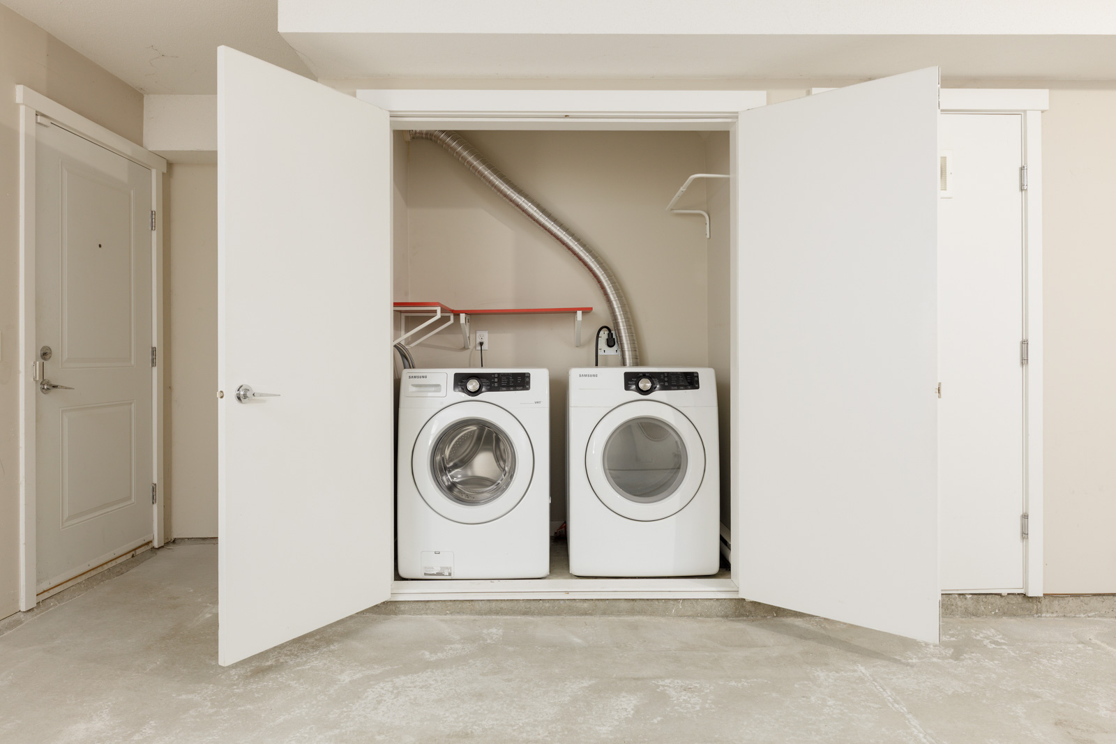 A laundry closet with open doors reveals a front-loading washer and dryer, a metal vent hose, a red hanging rod, and empty shelves above the appliances.