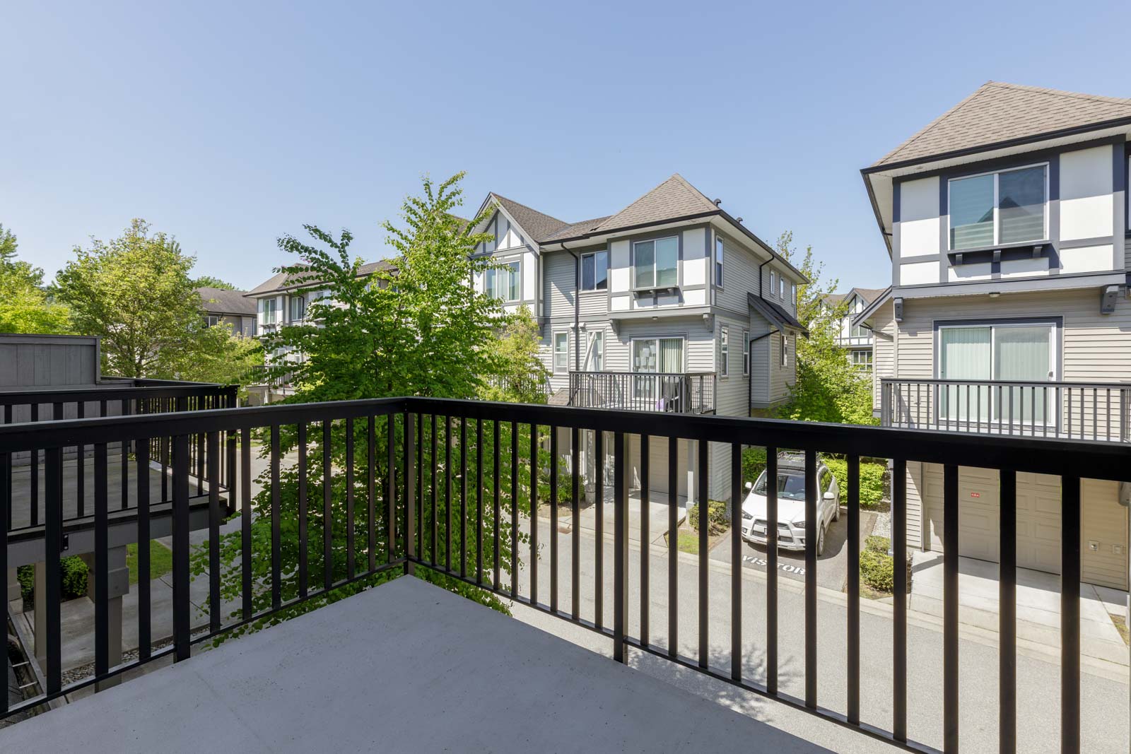 View from a balcony overlooking a residential street with modern townhouses, a parked car, and trees under a clear blue sky.