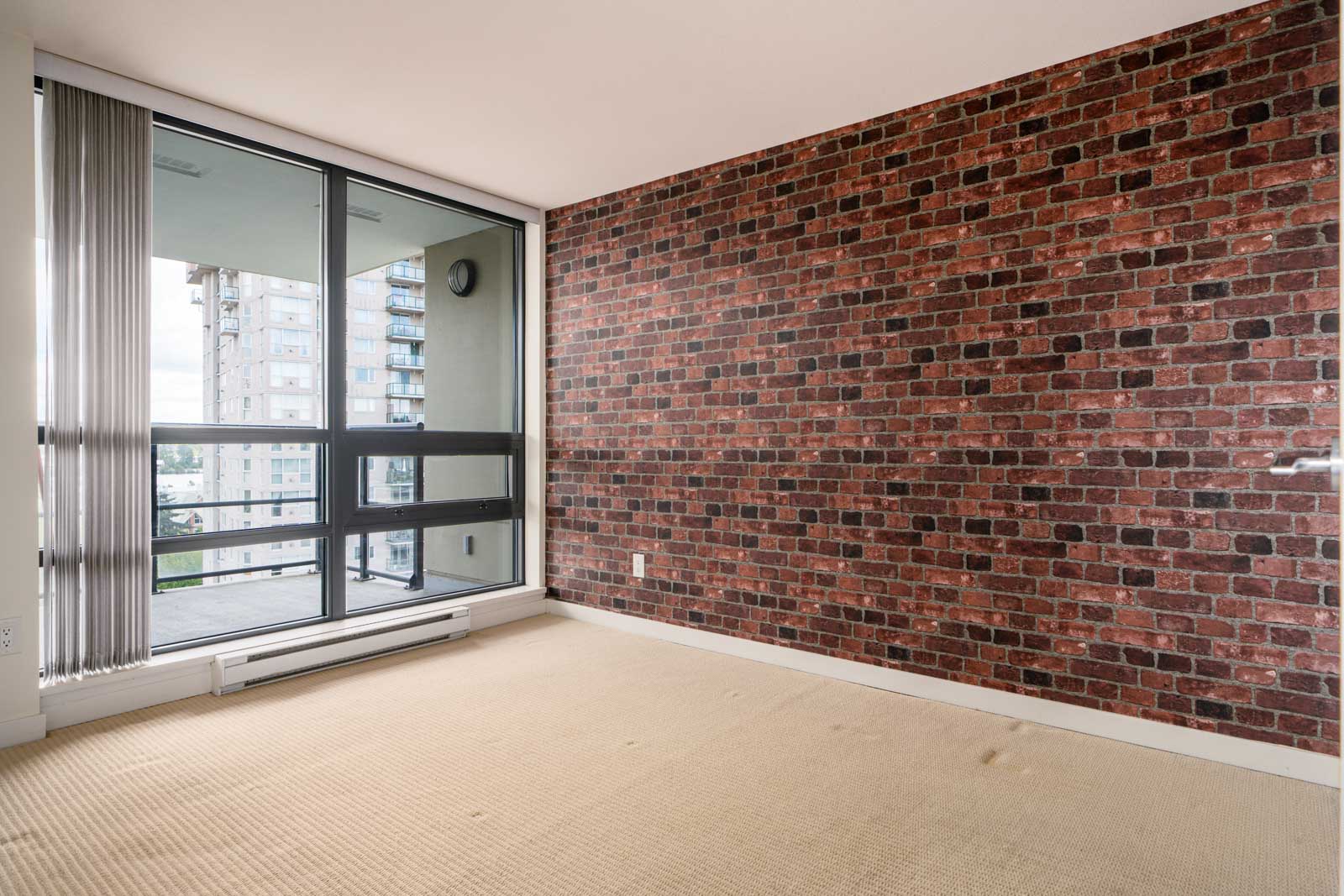 Empty room with a brick accent wall, beige carpet, and large sliding glass doors leading to a balcony with a view of nearby apartment buildings.
