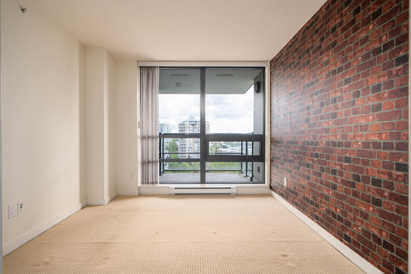 Unfurnished room with a large window and sliding glass door, an exposed brick accent wall, and beige carpet flooring. City buildings and greenery are visible outside.