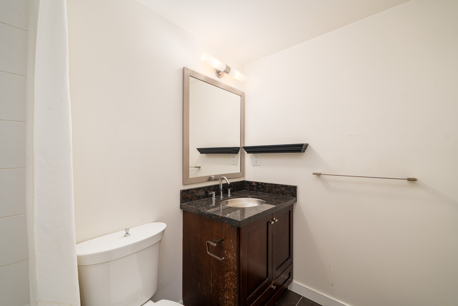 Small bathroom with a white toilet, dark wood vanity with sink, wall mirror, floating shelf, towel bar, and white walls.