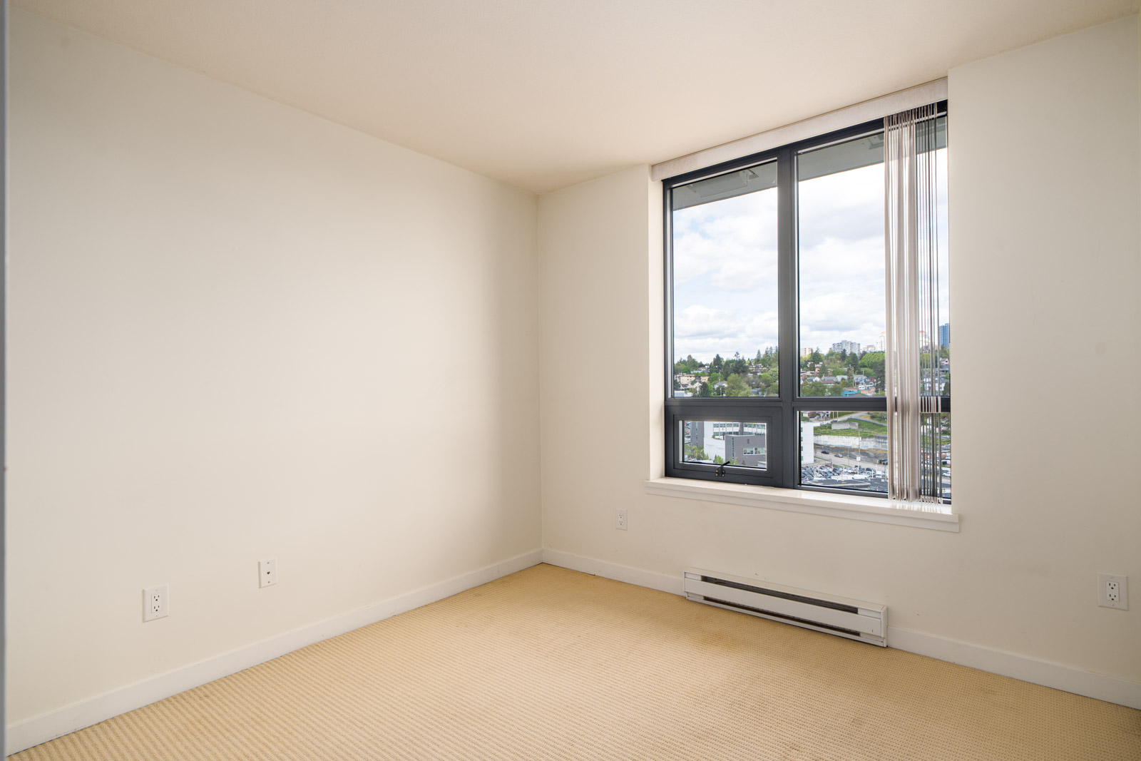 Empty room with beige carpet, off-white walls, and a large window showing an outdoor cityscape and cloudy sky.