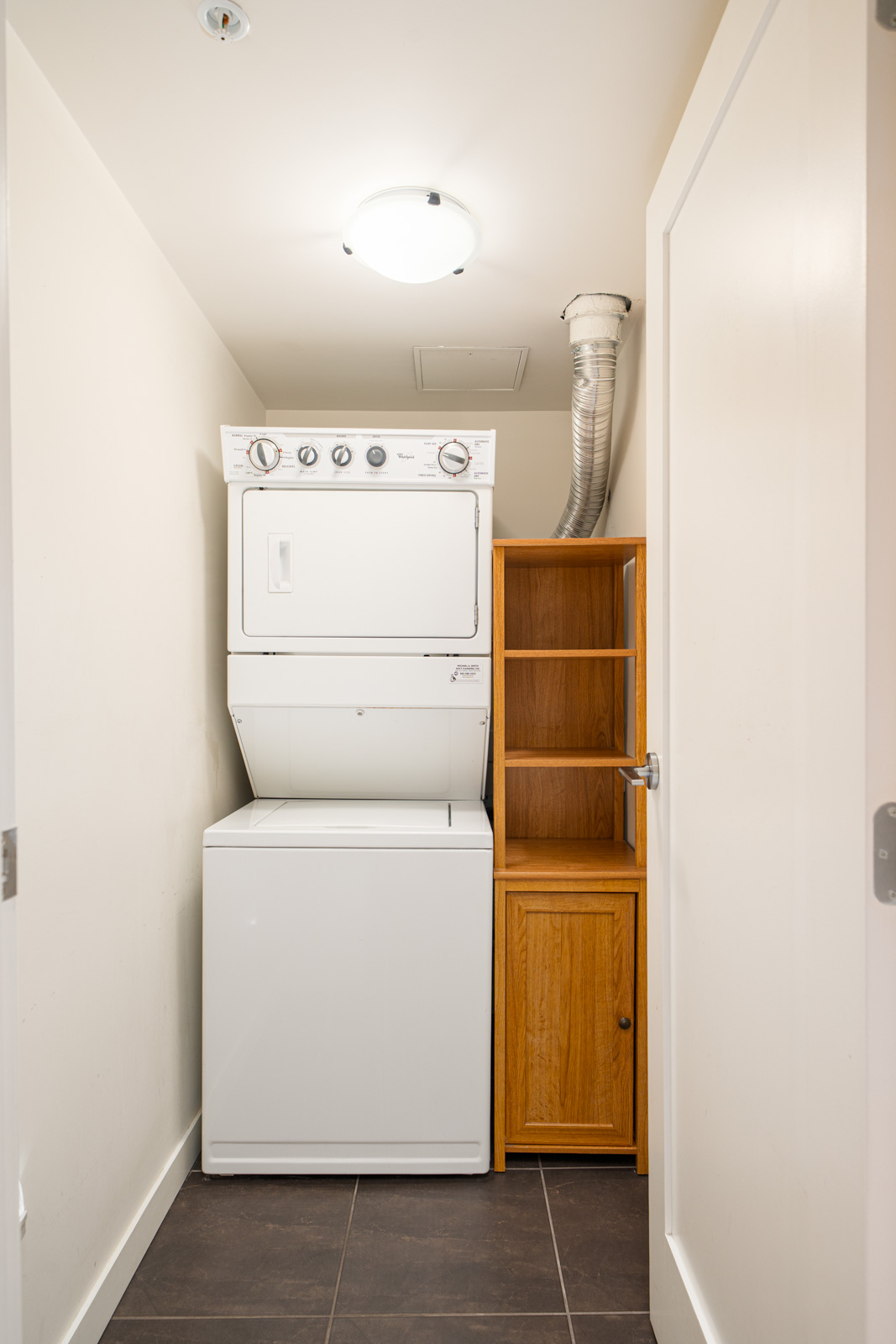 A small laundry room with a stacked washer and dryer next to a wooden shelving unit, white walls, and a tiled floor.