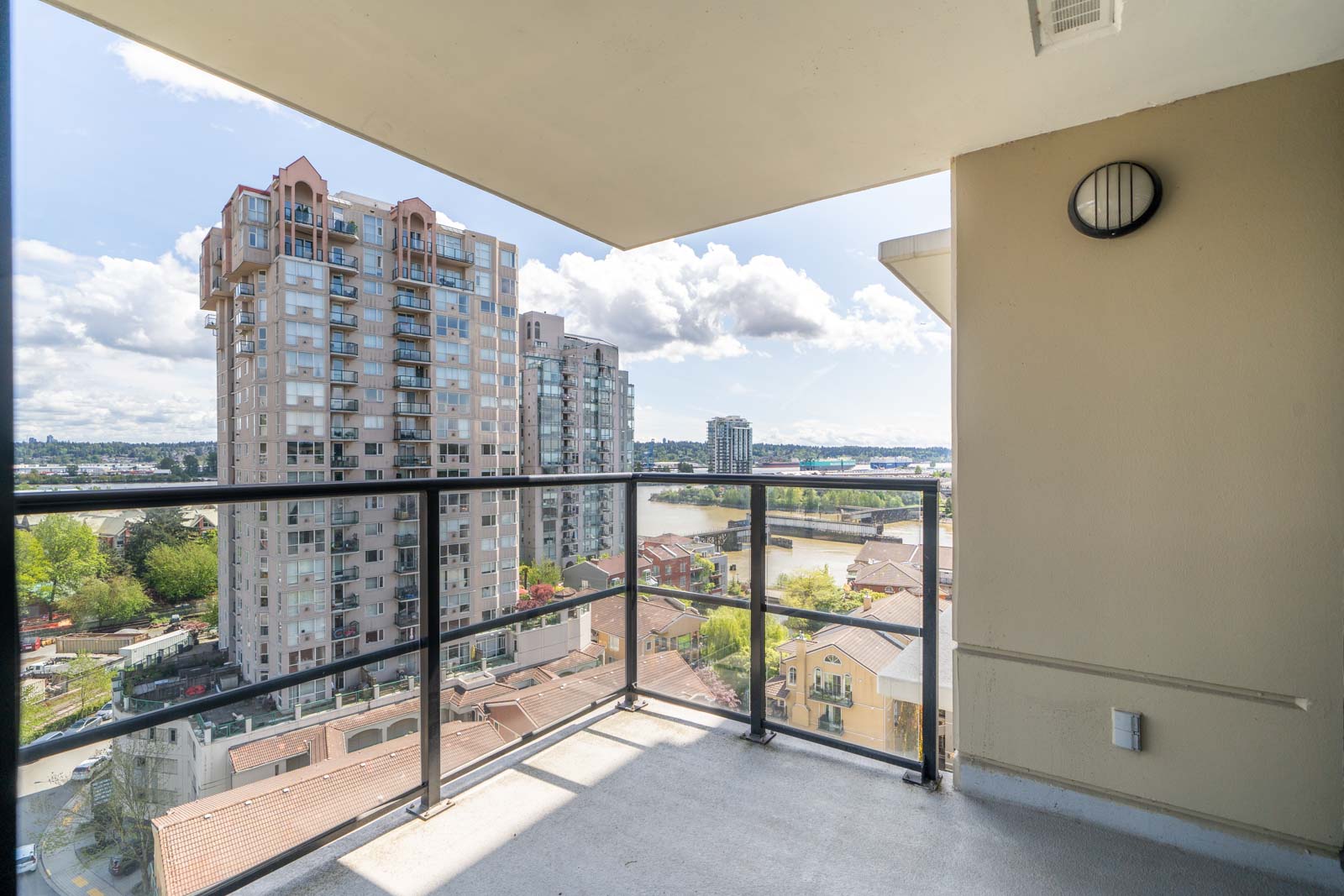 Balcony with glass railing overlooking high-rise buildings, a river, and a partially cloudy sky in an urban area.
