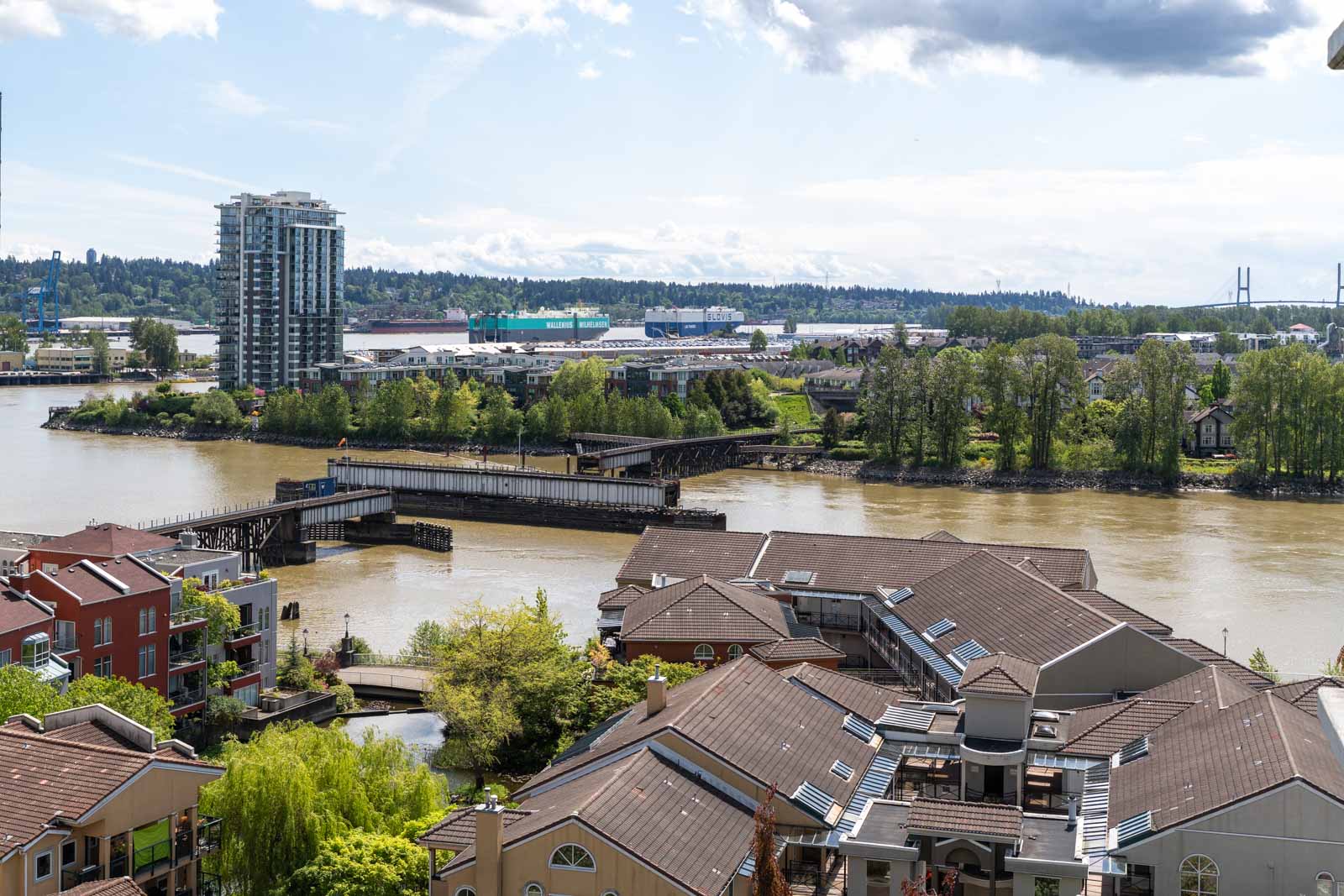 View of a river with residential buildings in the foreground, a bridge crossing the water, and tall modern buildings and trees in the background under a partly cloudy sky.