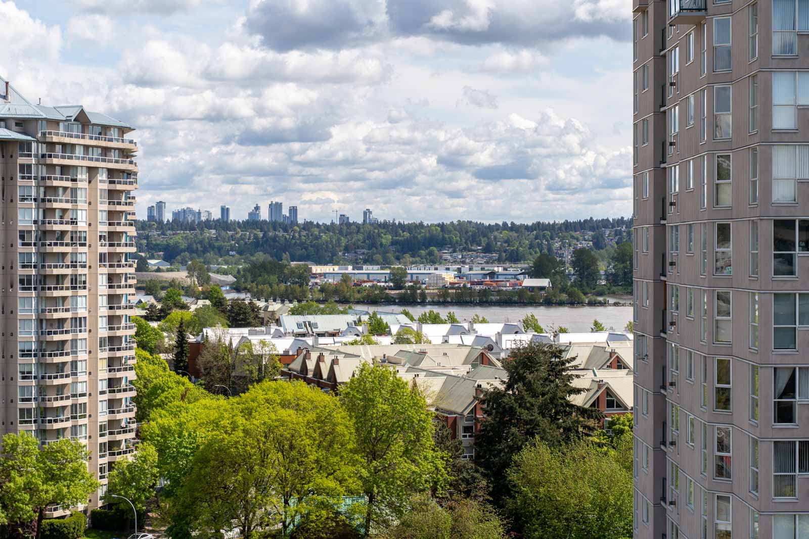 View of a cityscape with high-rise apartment buildings in the foreground, leafy trees below, and a river and distant skyline under a cloudy sky in the background.