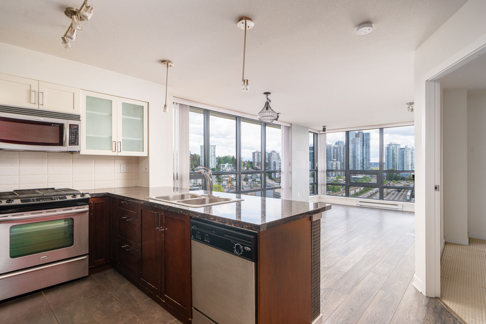 Modern apartment kitchen with stainless steel appliances, dark countertops, and an open layout leading to a bright living area with large windows overlooking a cityscape.