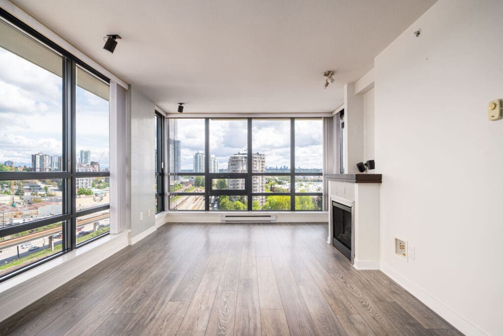 Unfurnished modern apartment living room with large floor-to-ceiling windows, wood flooring, and a small fireplace, offering a cityscape view.