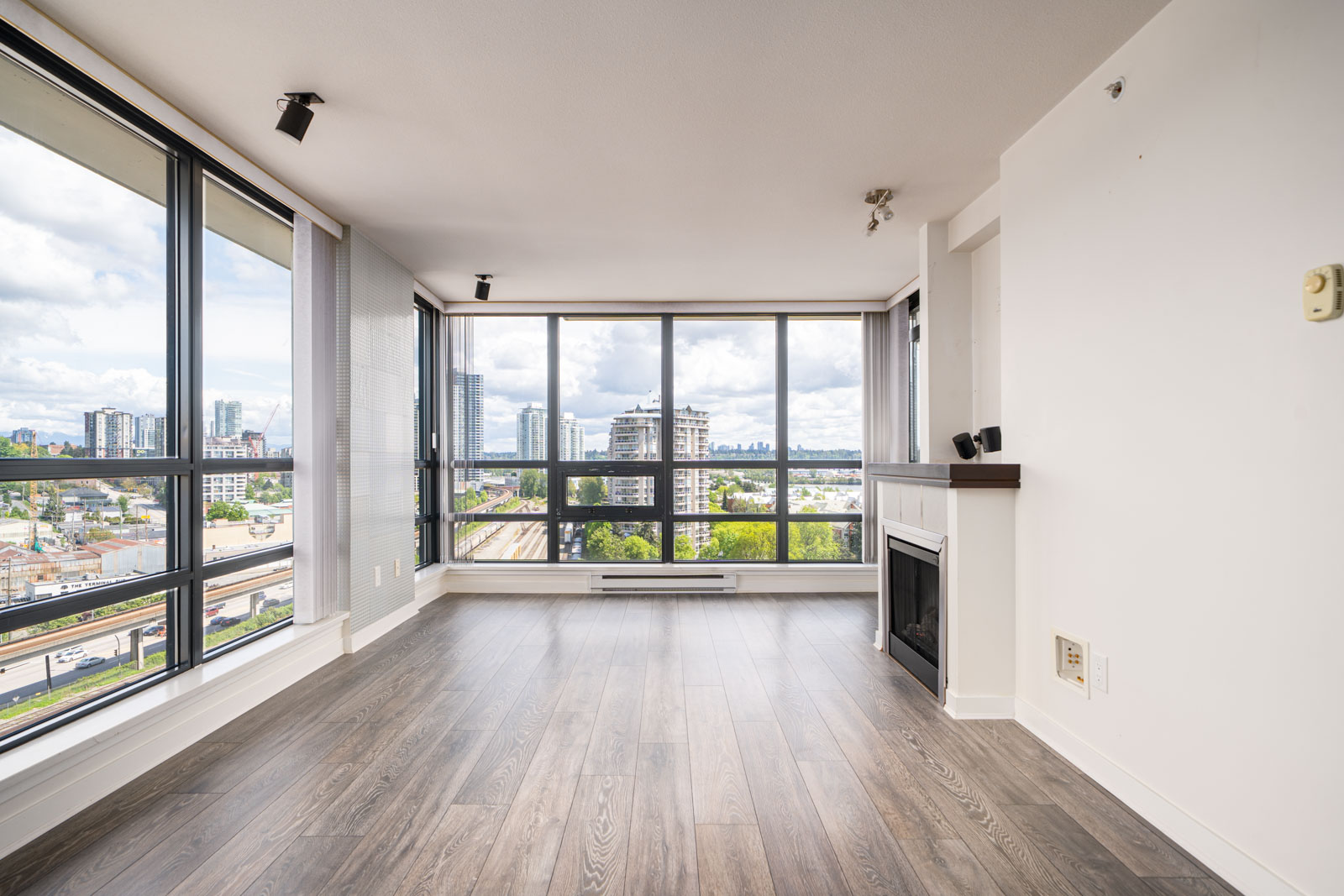 Unfurnished modern apartment living room with large floor-to-ceiling windows, wood flooring, and a small fireplace, offering a cityscape view.