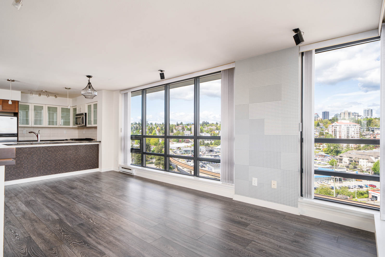 Open-plan modern condo with large windows, dark wood floors, and a view of the city skyline; kitchen area visible in the background.