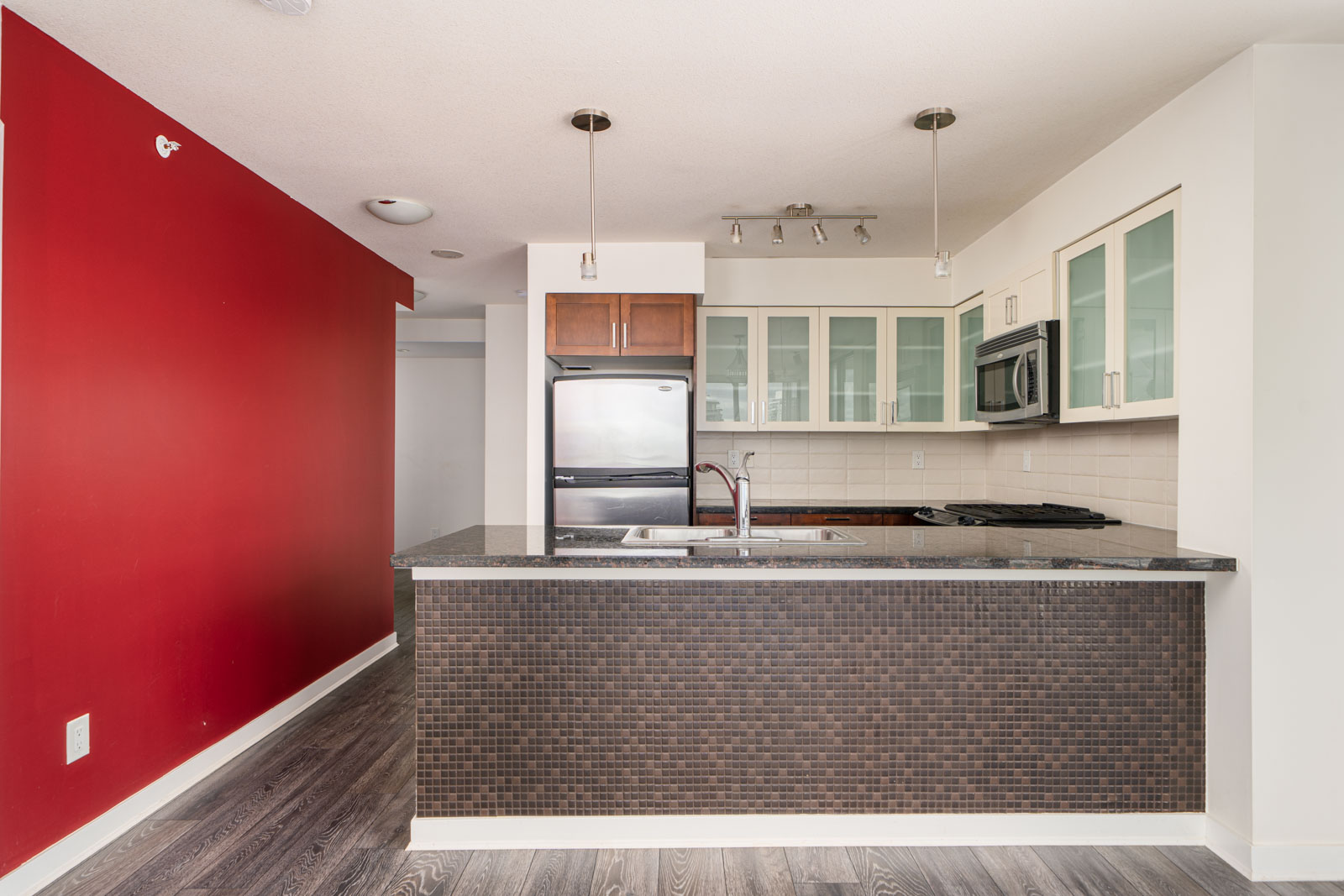 Modern kitchen with a black countertop, mosaic tile island, stainless steel appliances, glass-front cabinets, and a bold red accent wall.