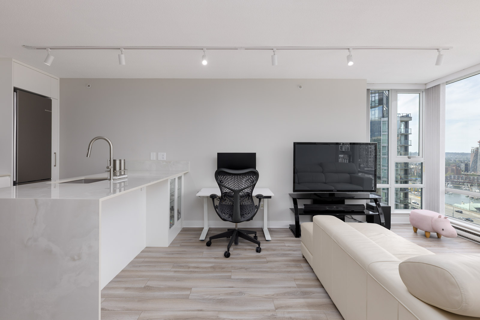 Modern apartment interior with a white kitchen island, ergonomic office chair at a desk, large TV, beige sectional sofa, and city view through floor-to-ceiling windows.