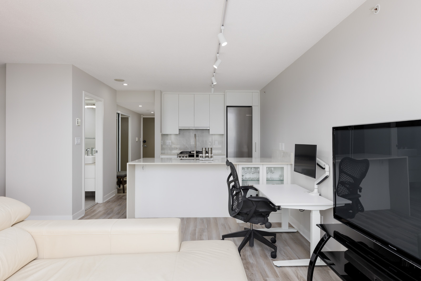 Modern open-plan apartment with a kitchen, home office setup with a desk and chair, cream sofa, and a large flat-screen TV on a stand.