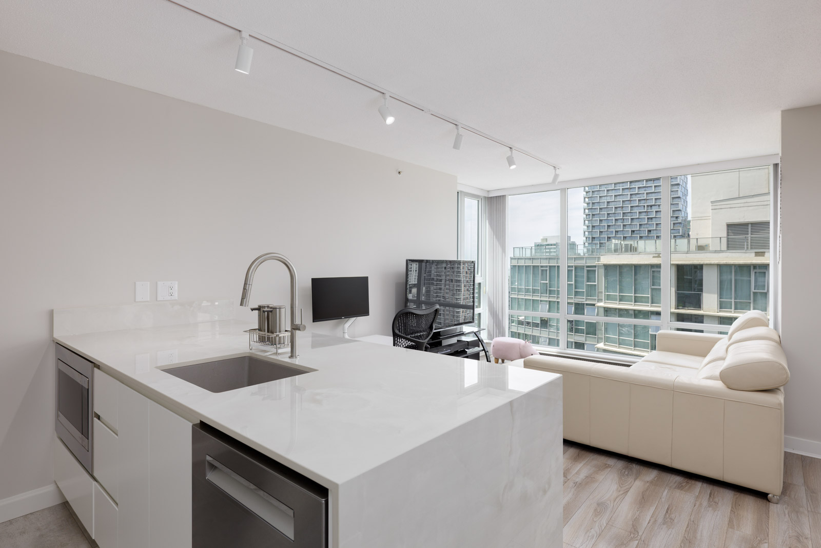 Modern apartment interior with a white kitchen island, sink, built-in appliances, a beige sofa, desk setup, and large windows overlooking city buildings.