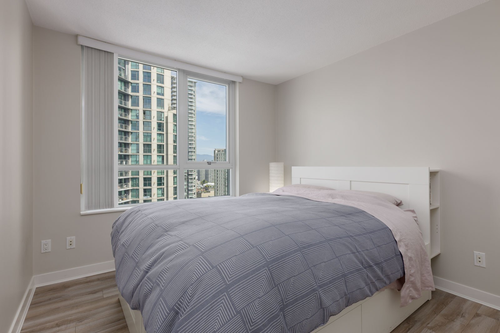 Modern bedroom with a large window showing city buildings, a bed with a gray patterned comforter, and light-colored walls and flooring.