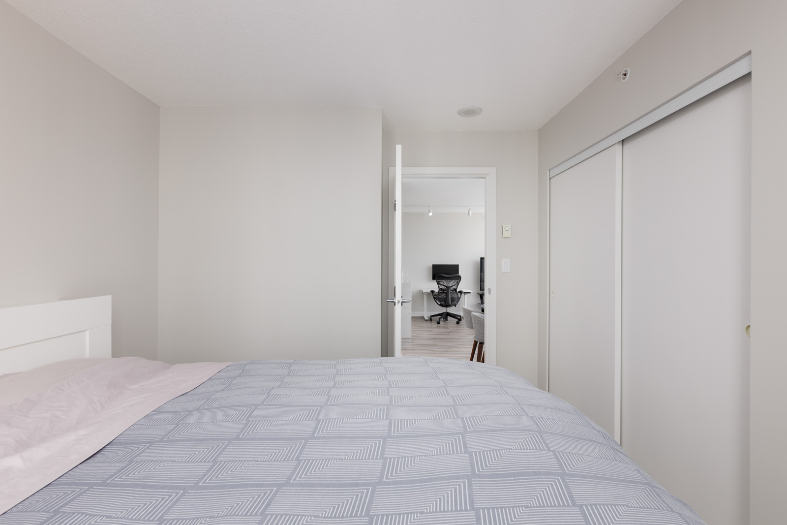 Bedroom with a made bed in the foreground, white walls, closed closet doors, and an open door revealing a home office with a chair and desk.